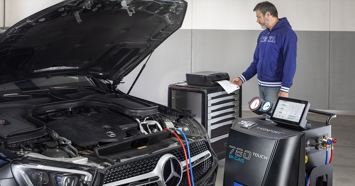 ac-diagnostics-featured-konfort-780-1200×630-1 TEXA KONFORT 780 Touch Bi-Gas A/C service machine with its touchscreen on, connected by red and blue hoses to an SUV with the hood open in a shop, as a technician reviews a printed report.