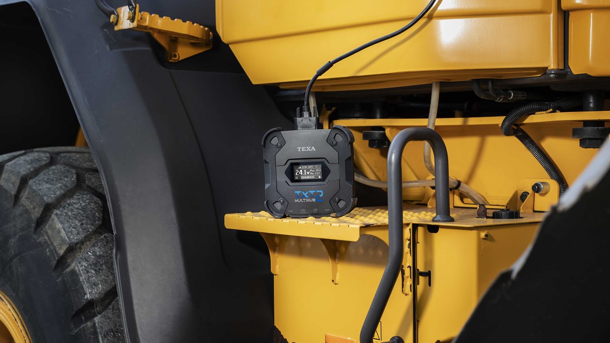 TEXA Navigator TXT Multihub 2 with its display on, reading 24.1 V in diagnosis mode, sitting on the step of a yellow wheel loader and cabled to the machine.